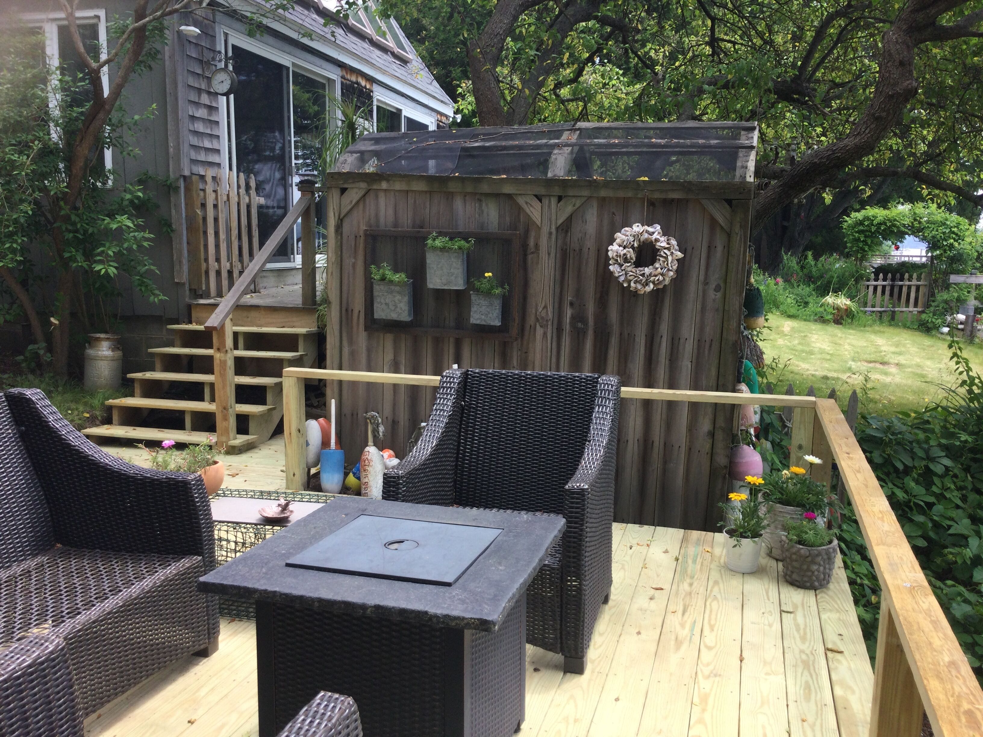 Una terraza con mobiliario de mimbre y una pequeña mesa, una caseta de madera con un adorno y una escalera que lleva a una casa.