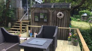 Una terraza con mobiliario de mimbre y una pequeña mesa, una caseta de madera con un adorno y una escalera que lleva a una casa.