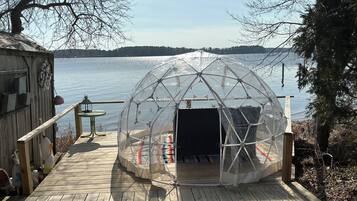A transparent geodesic dome structure on a wooden deck by a lake.