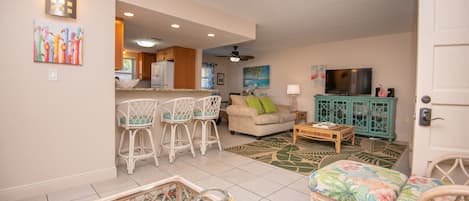 A living room with a sofa, a coffee table, and a TV. There is a kitchen area with a refrigerator and a dining table with chairs.