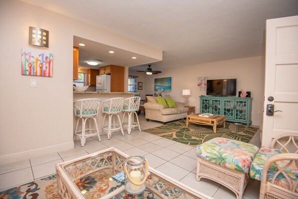 A living room with a sofa, a coffee table, and a TV. There is a kitchen area with a refrigerator and a dining table with chairs. - Walk to Beach | Parking Space Provided (St. Pete Beach)