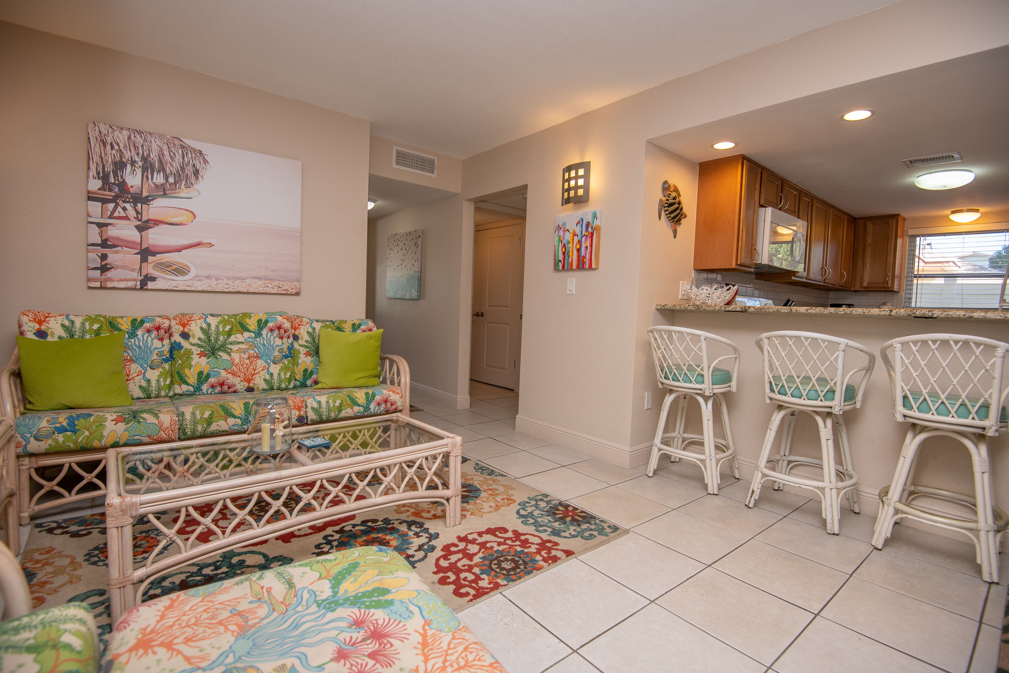 A living room with a colorful sofa, a wicker coffee table, and a kitchen area with white bar stools.