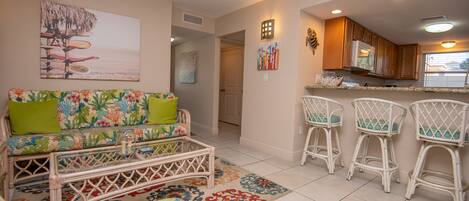 A living room with a colorful sofa, a wicker coffee table, and a kitchen area with white bar stools.