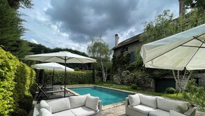 Un espace aménagé au bord de la piscine, avec des meubles d’extérieur blancs et des parasols, entouré de verdure et bordé d’un bâtiment en pierre.