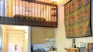 Eine Küche mit Kochinsel, ein Essbereich mit Stühlen, ein Schlafzimmer im Zwischengeschoss mit Balkon und eine große Wandteppich.