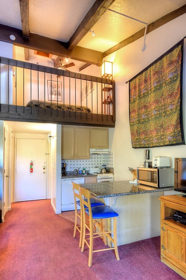 A kitchen with a central island, a dining area with chairs, a loft bedroom with a balcony, and a large tapestry on the wall.