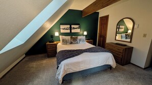 A bedroom with a sloped ceiling, a bed with white bedding, a dark blue blanket, two bedside tables with lamps, a dresser, a mirror, and a framed picture on the wall.