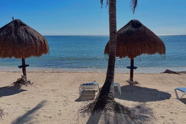 A beach with two thatched umbrellas and lounge chairs facing the ocean.