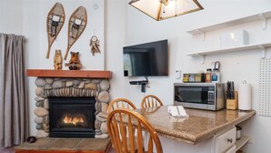 A cozy living room with a stone fireplace, a flat-screen TV, a dining area with wooden chairs, and a kitchen with a microwave and storage shelves. - (B210) Loft Condominiums (Yosemite National Park)