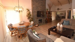 A cozy living room with a stone fireplace, a dining area with wicker furniture, and a modern kitchen in the background.