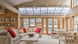 A spacious living room with a large sectional sofa, a coffee table, and a dining area with a table and chairs. There are large windows with a view of snow-covered trees. - Ski-In/Ski-Out Winter Park Condo w/ Pool Access! (Winter Park)