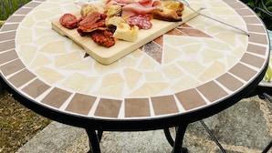 A round table with a mosaic pattern, a charcuterie board with various meats and cheeses, and two glasses of white wine, set outdoors with a view of rolling hills and a tall tree in the background. - Beautiful house near San Gimignano in Tuscany with views, pool,garden, A/C, WiFi (Gambassi Terme, FI)