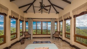A spacious bedroom with a large bed, two chairs, a ceiling fan, and large windows offering a view of the landscape. - Platinum - A New level of Luxury, with 360 degree views & unmatched quality (Quepos)