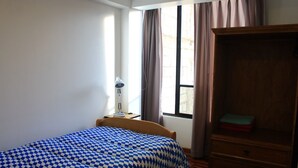 Un dormitori amb un llençol de sobreposició de quadres, un armari de fusta, un escriptori amb una làmpada i una finestra amb cortines.
