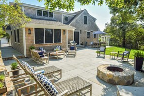 Un patio techado con mobiliario exterior, una chimenea y vista a una casa al fondo.