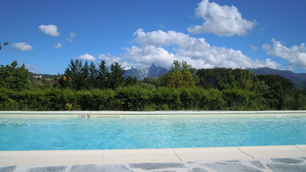 A swimming pool with clear blue water, surrounded by stone tiles. In the background, there are trees and mountains under a blue sky with clouds. - Podere la Piana-Cedro (fivizzano)
