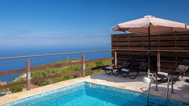 A poolside area with a view of the sea, wooden deck, lounge chairs, and an umbrella.