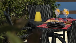 An outdoor dining setup with wicker chairs, a table with a red checkered tablecloth, two glasses of a clear beverage with orange slices, a bottle of orange juice, and strawberries.