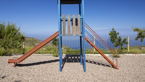 A playground structure with a slide and climbing area, surrounded by a fence and gravel ground.