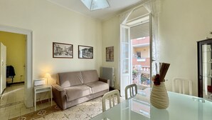 A living room with a sofa, a glass table, and a vase with flowers. There are framed pictures on the wall and a window with a view of buildings outside. - Da Zio Giovanni - 2 steps from the sea (Pescara)