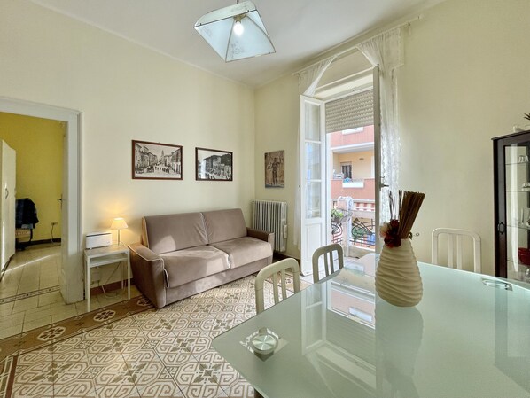 Una sala de estar con un sofá, una mesa de vidrio y un jarrón con flores. Hay cuadros en la pared y una ventana que ofrece vistas de edificios.