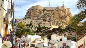 Un pueblo costero con una colina rocosa destacada al fondo, edificios blancos con balcones y un cartel que indica un negocio local.
