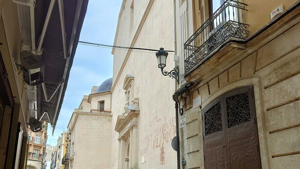 Un café al aire libre con mesas y sillas, frente a un edificio con balcones ornamentados y una gran puerta de madera.