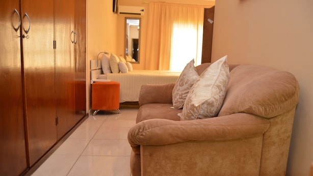 Een woonkamer met een beige bank, twee kussens, een klein oranje pouf en een bed met een witte deken en kussens.