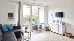 Ein Wohnzimmer mit grauem Sofa, einem kleinen Korbstuhl mit Pflanze, einem weißen Schrank mit Fernseher und einem großen Fenster mit Blick ins Grüne.