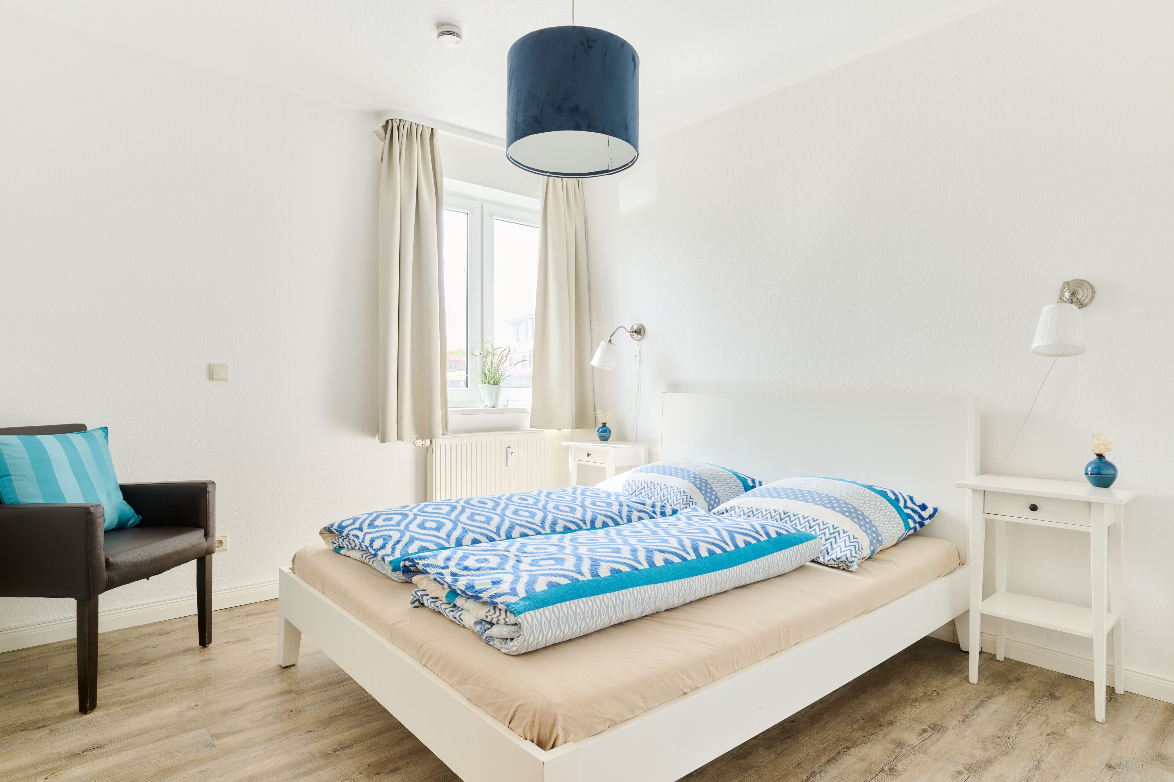 Un dormitorio moderno con una cama blanca, una lámpara colgante azul, una mesita de noche blanca, una silla y una ventana con cortinas.