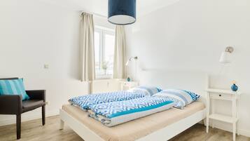 Un dormitorio moderno con una cama blanca, una lámpara colgante azul, una mesita de noche blanca, una silla y una ventana con cortinas.