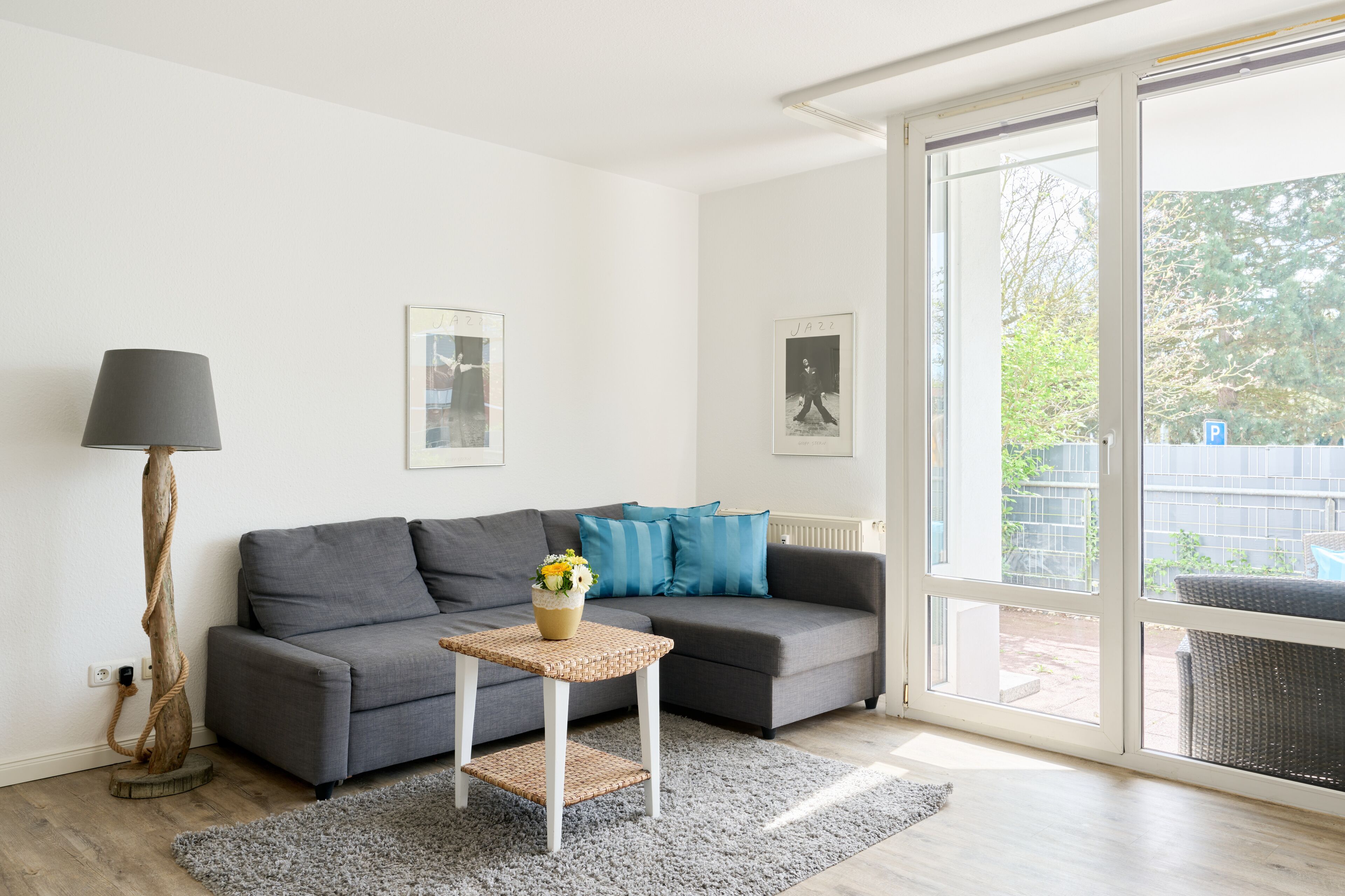 Una sala de estar moderna con un sofá gris, una mesita con un jarrón, una lámpara de pie y un amplio ventanal corredera que da a un espacio exterior.