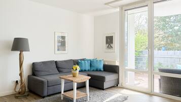 Una sala de estar moderna con un sofá gris, una mesita con un jarrón, una lámpara de pie y un amplio ventanal corredera que da a un espacio exterior.