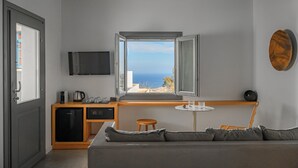A modern room with a large window, a sofa, a small table, a TV, and a kitchenette. - Lemon Suites Santorini (Santorini)