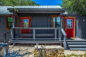 A small, rustic cabin with a metal roof, a wooden deck, and a red door.