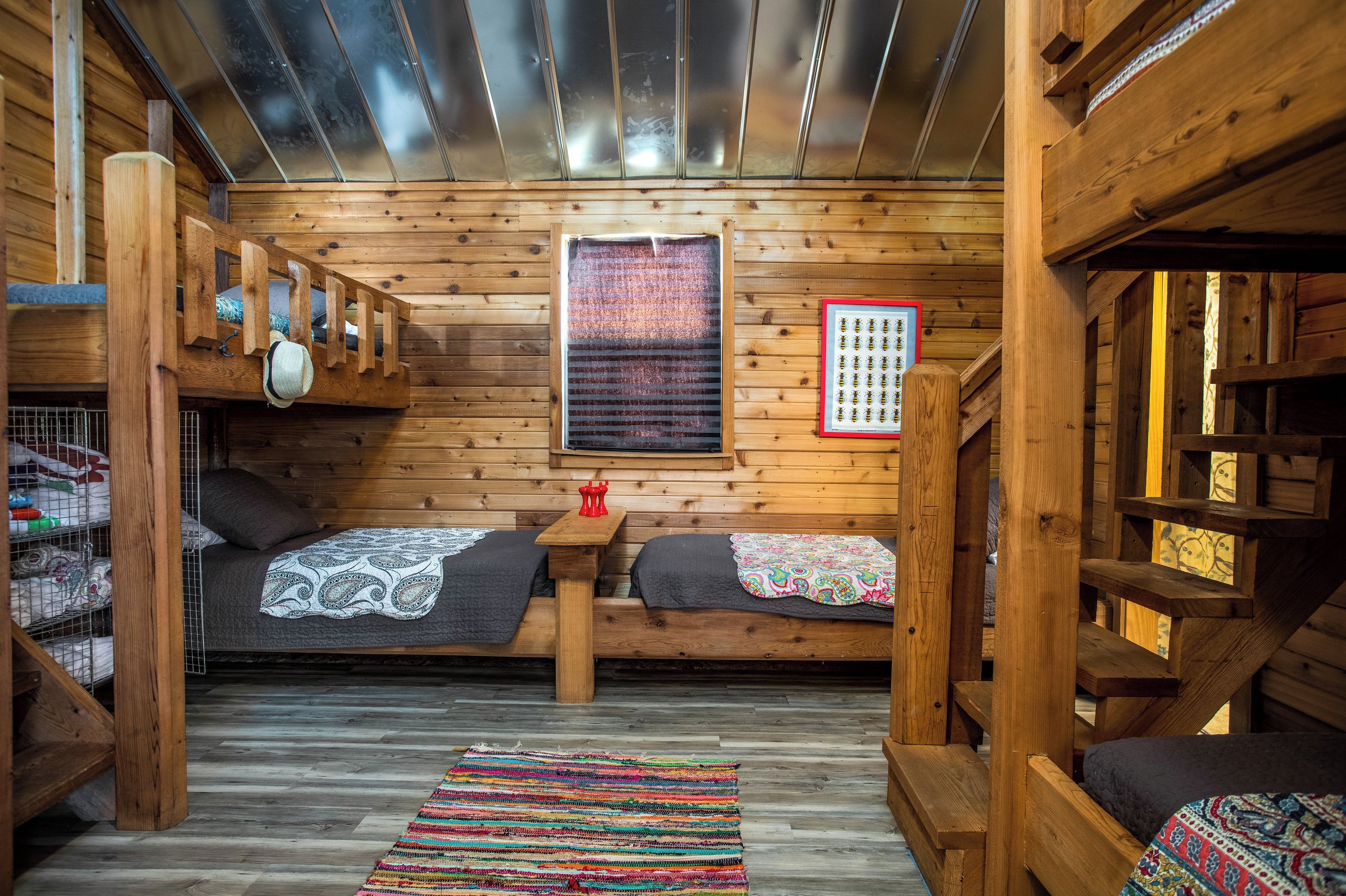 A cabin interior with wooden walls, a ceiling with metal panels, two beds with headboards, a small table, and a colorful woven rug.