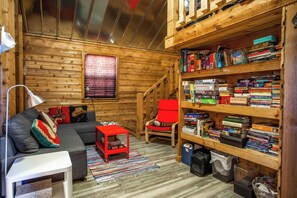 A cozy cabin interior with a wooden interior, a bookshelf filled with books, a red chair, a gray sofa, and a small table.