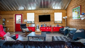 Interior acollidor d'una cabana, amb elements de fusta, una porta vermella, una televisió plana gran i una zona de seients confortable amb sofà i cadires.