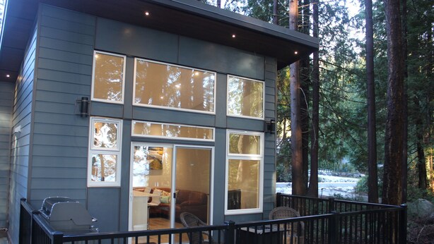 A modern cabin with large windows, a deck, and a view of the surrounding forest.