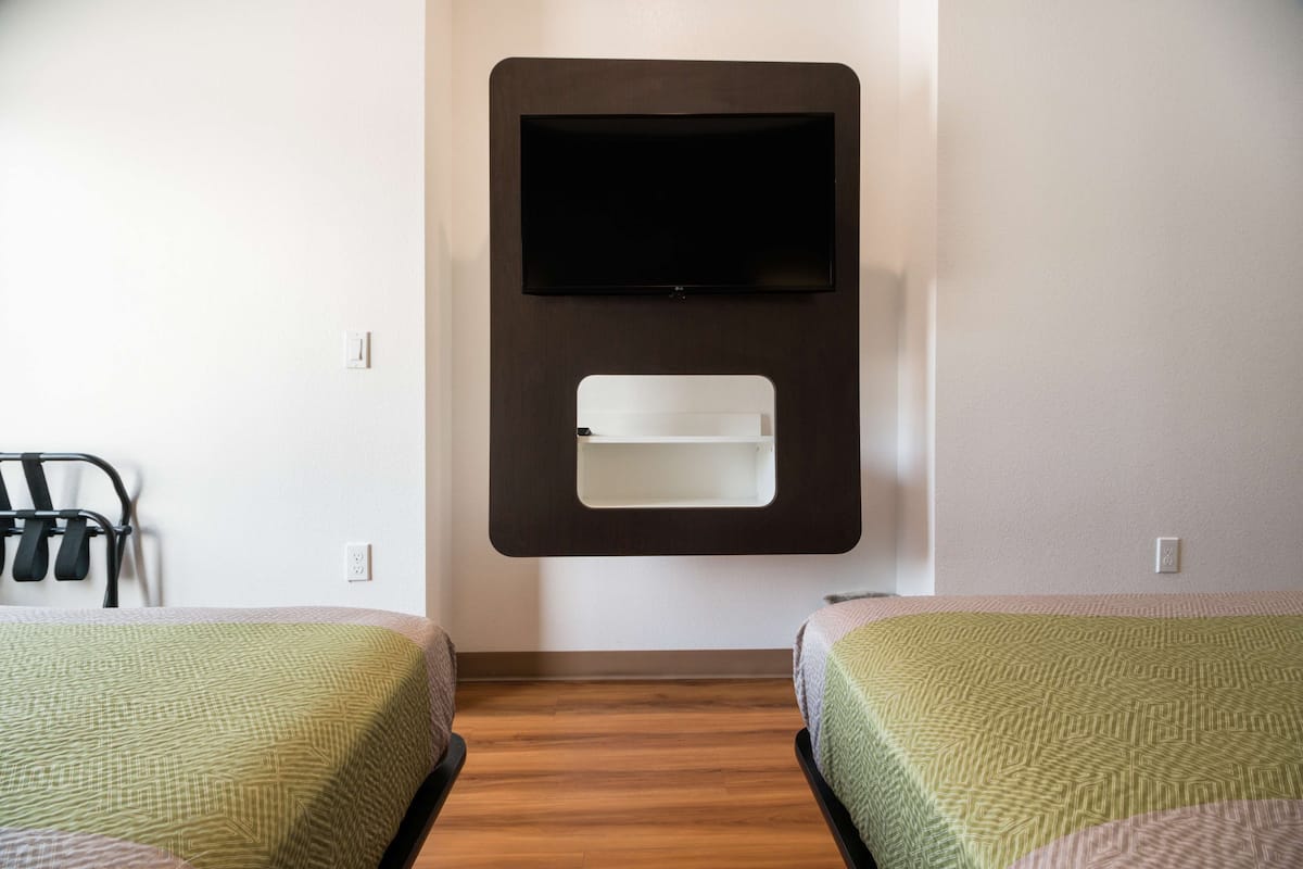 A hotel room with two beds, a mounted TV, and a small shelf.