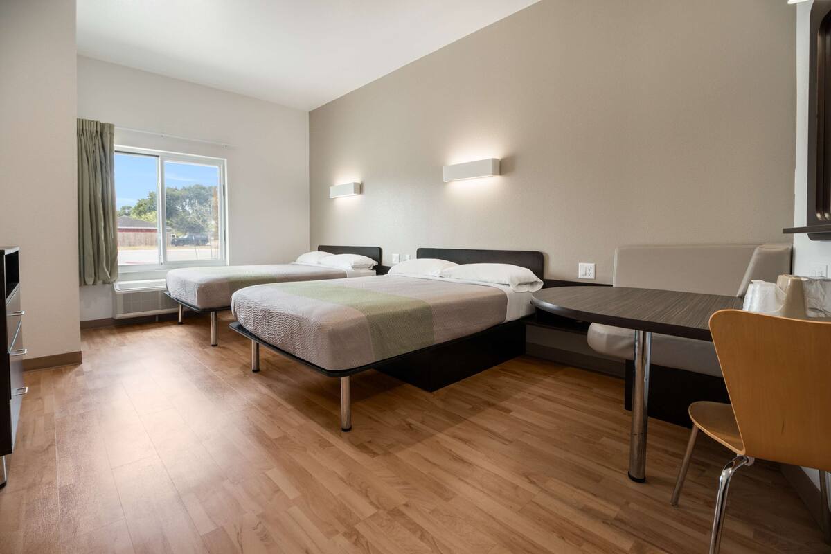 A hotel room with two beds, a dining table, a chair, and a window with curtains.