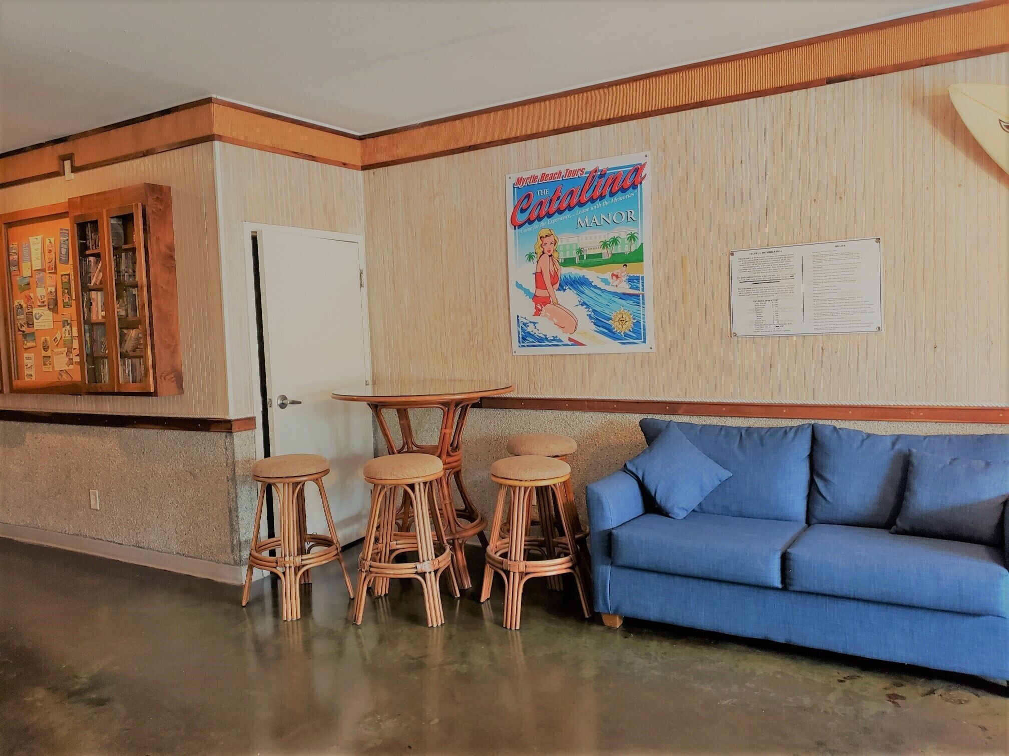 A room with a blue sofa, wicker chairs, a wooden table, a door, and a wall with a poster and a menu.