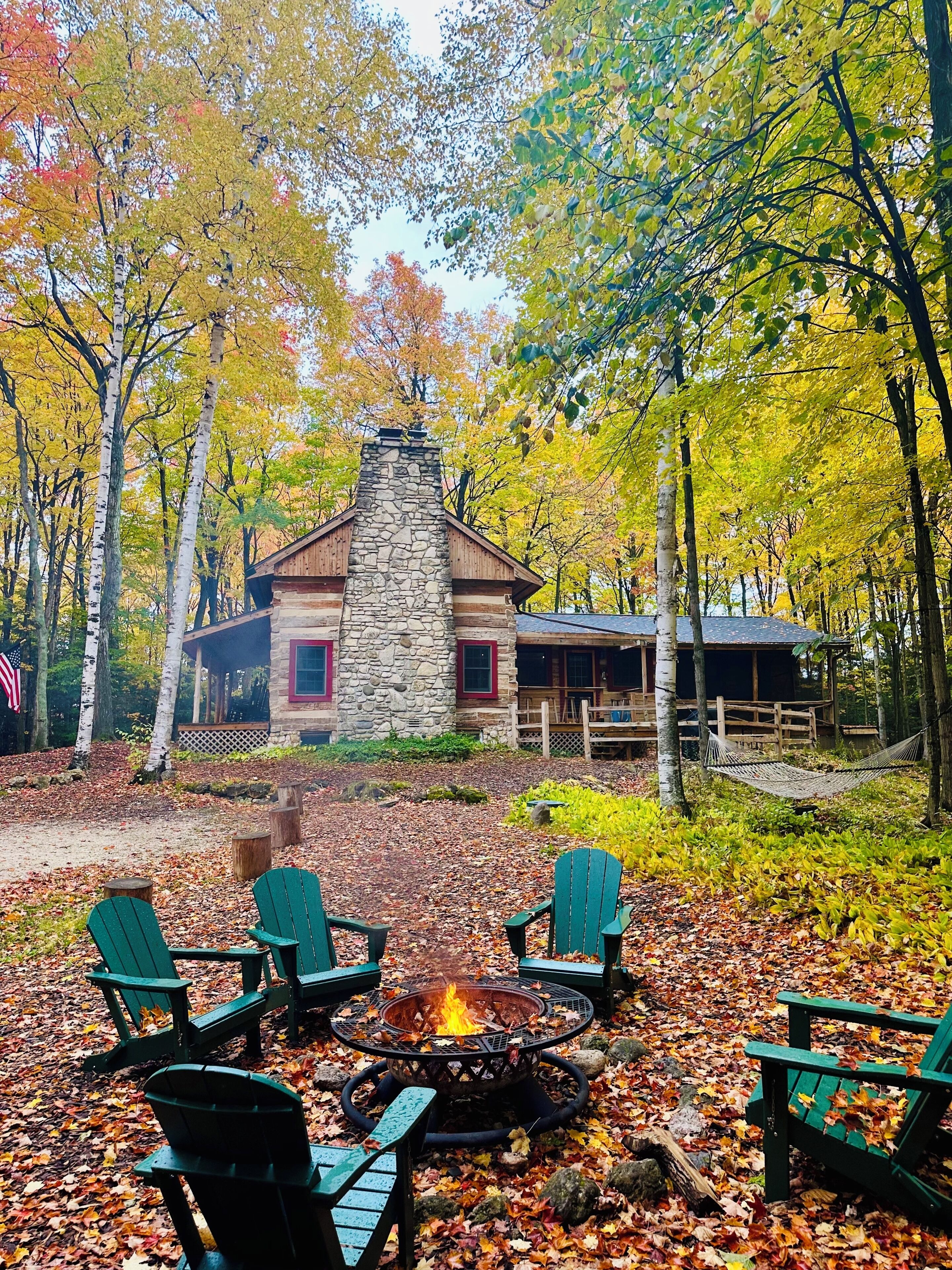 Un chalet doté d’une cheminée en pierre, d’un foyer et de fauteuils Adirondack verts.