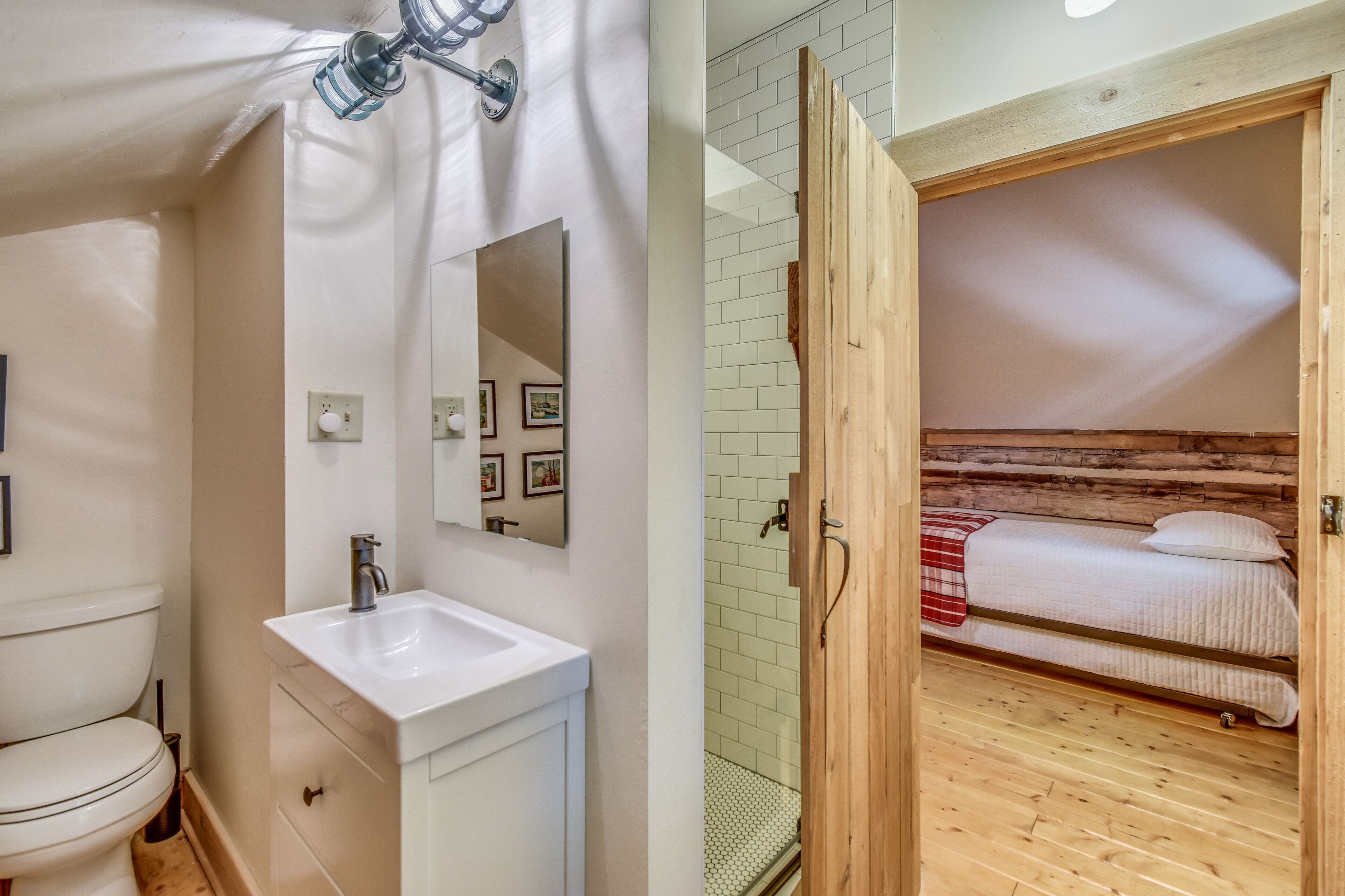 Une salle de bain avec un lavabo blanc, un miroir et des toilettes. La pièce est dotée d’un plancher en bois et d’une porte en bois qui donne sur une chambre à coucher avec un lit et une couverture à carreaux rouges.