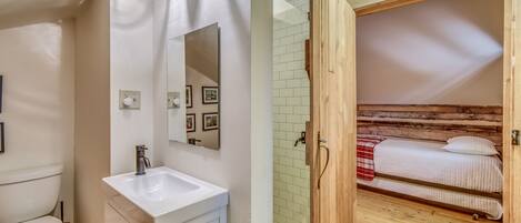Une salle de bain avec un lavabo blanc, un miroir et des toilettes. La pièce est dotée d’un plancher en bois et d’une porte en bois qui donne sur une chambre à coucher avec un lit et une couverture à carreaux rouges.