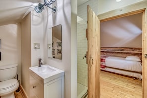 Un baño con lavamanos blanco, espejo y inodoro. El piso es de madera y una puerta de madera da paso a un dormitorio con cama y una manta roja a cuadros.