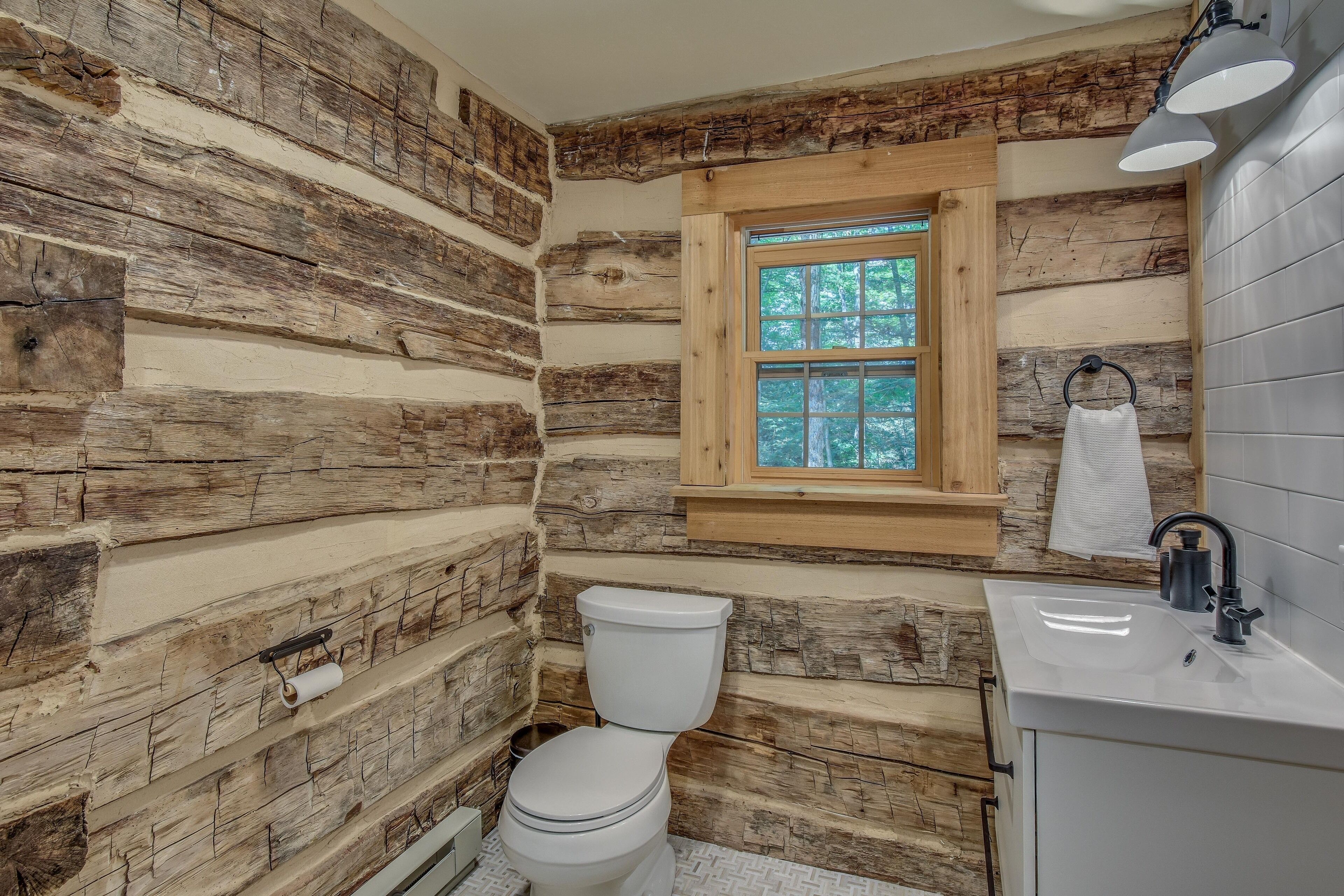 A rustic cabin bathroom with a wooden log wall, a window, a toilet, and a sink.