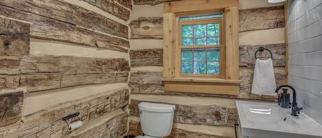 A rustic cabin bathroom with a wooden log wall, a window, a toilet, and a sink.