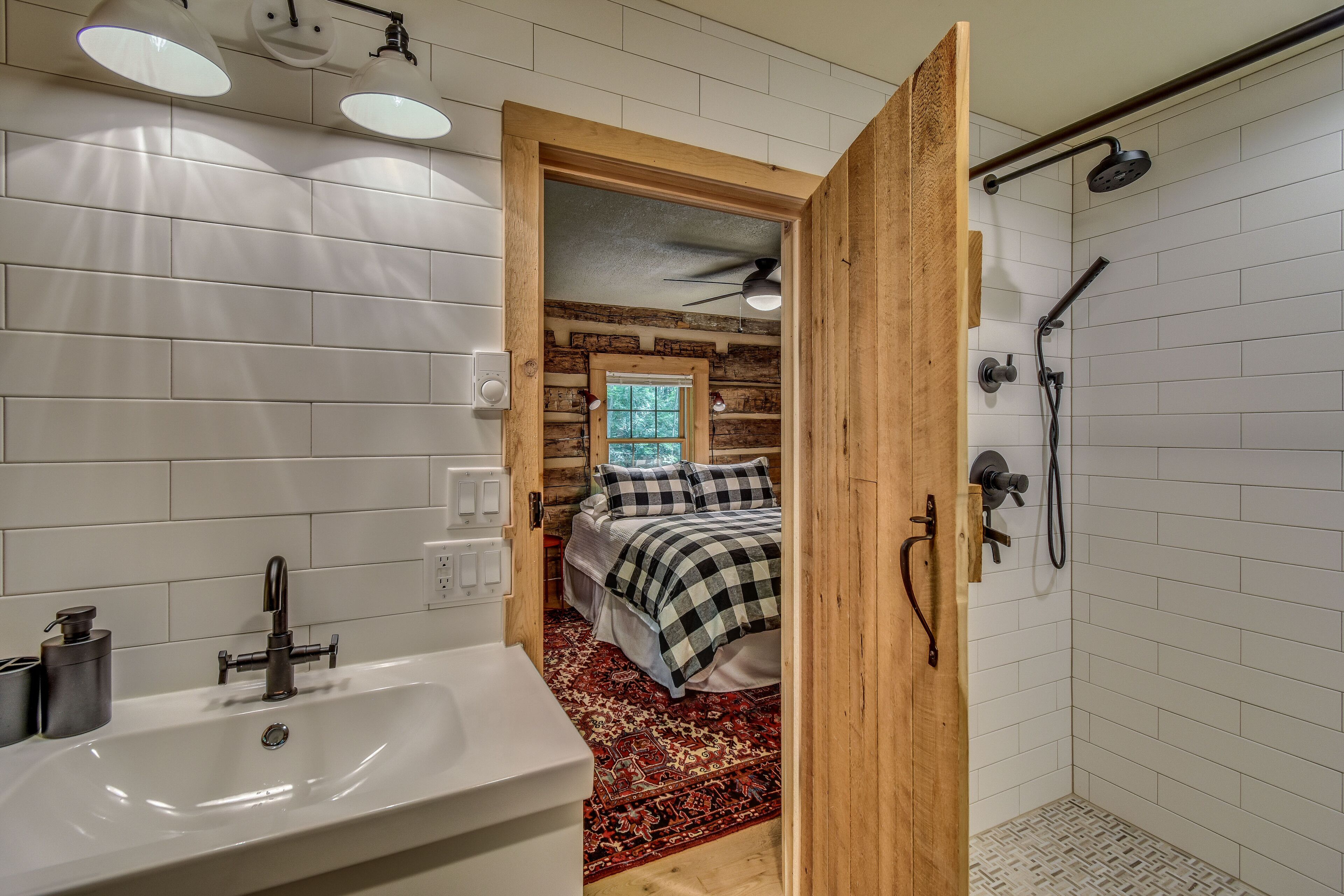 A bathroom with a sink, shower, and a wooden door leading to a bedroom with a bed and a window.