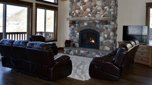A living room with a stone fireplace, leather sofas, a flat-screen TV, and large windows. - Yellowstone Northern Views -- Gorgeous Home. One mile to Yellowstone! (Gardiner)